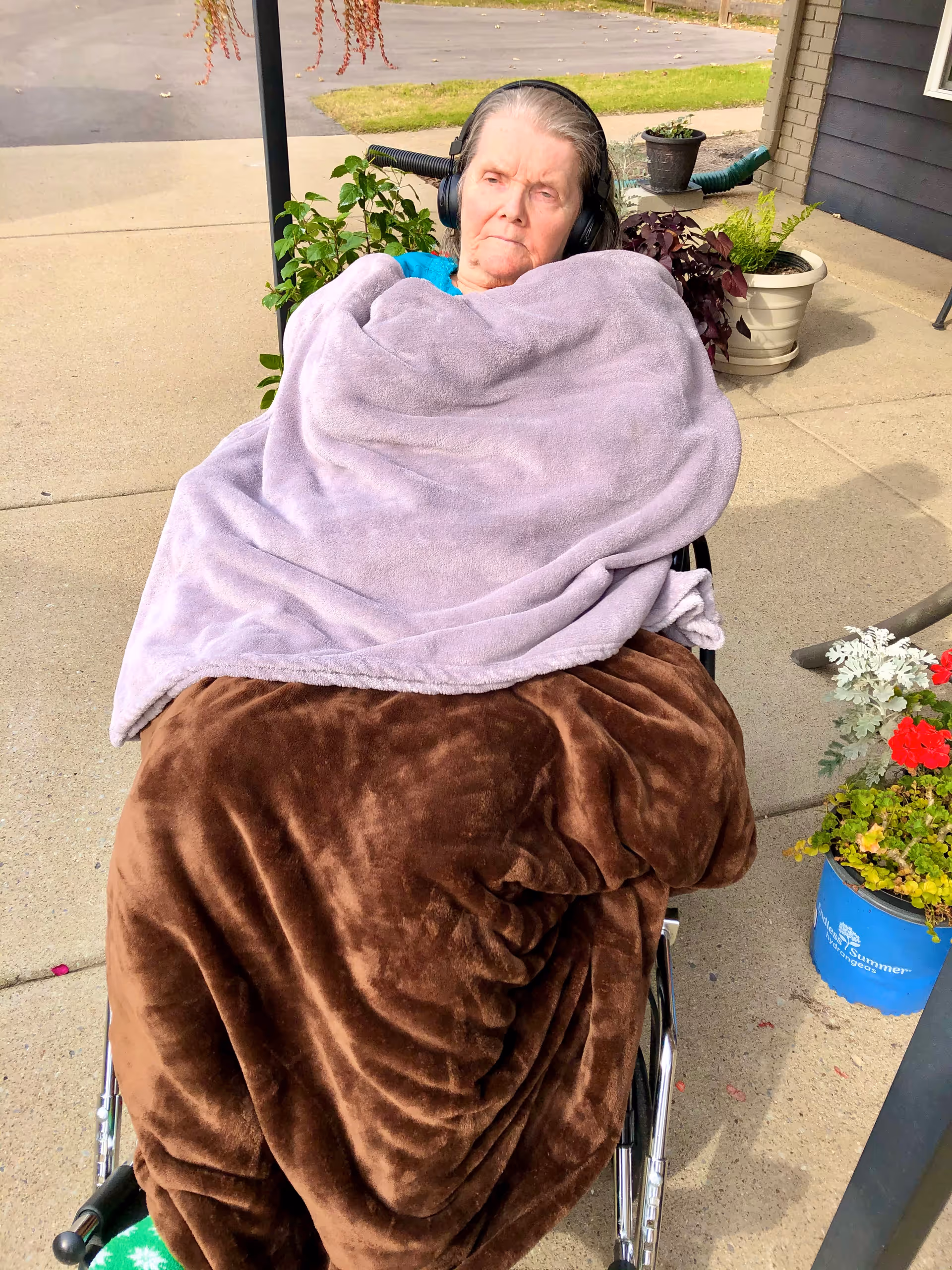 An elderly woman sitting in a wheelchair outdoors, wrapped in a brown blanket and a lavender blanket, wearing headphones. There are potted plants and a building exterior visible in the background.