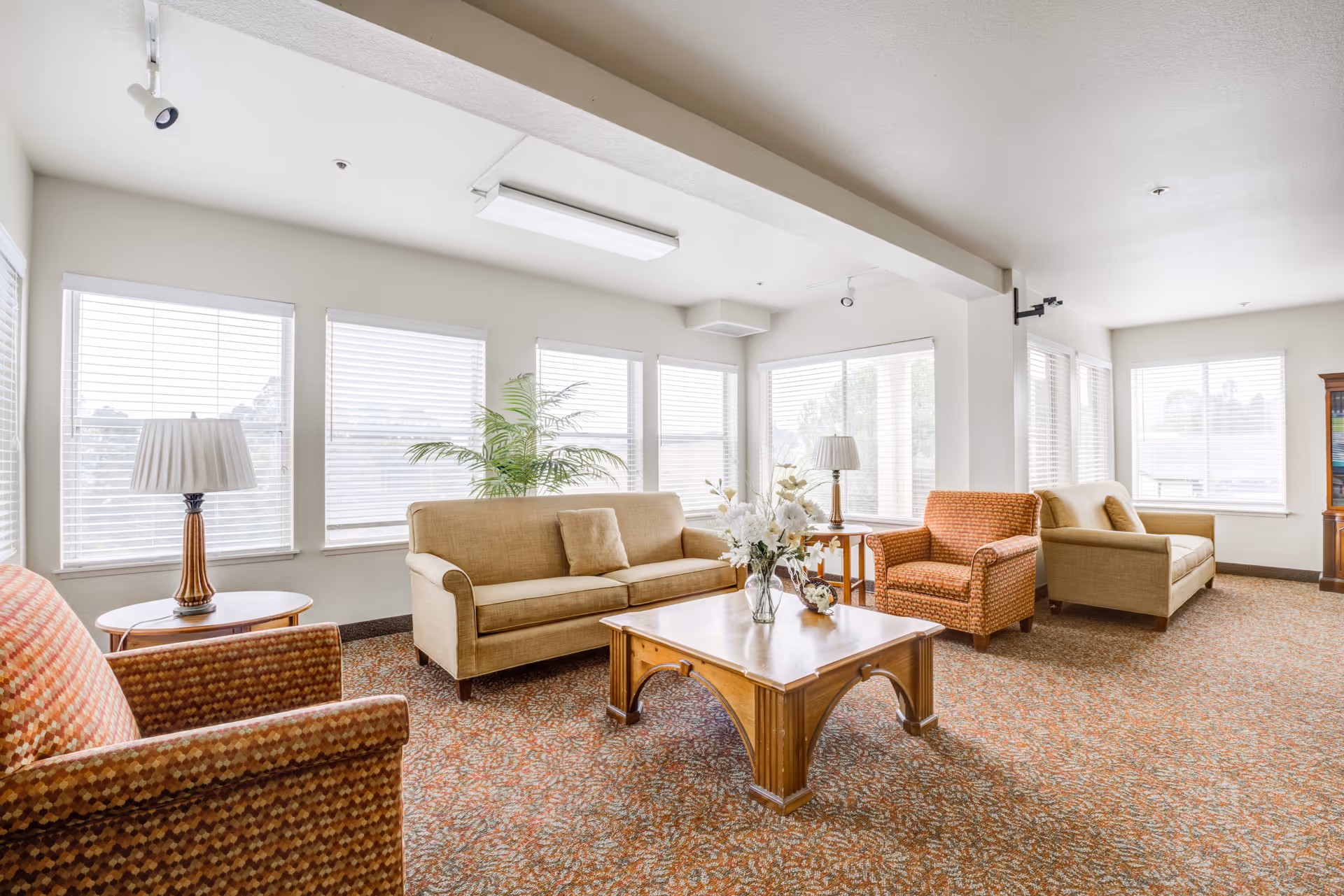 A bright and spacious living room area with large windows covered by white blinds. The room features two beige sofas, two patterned armchairs, a wooden coffee table with a floral arrangement, two side tables with lamps, and a potted plant near the windows. The carpet has a patterned design in warm tones.