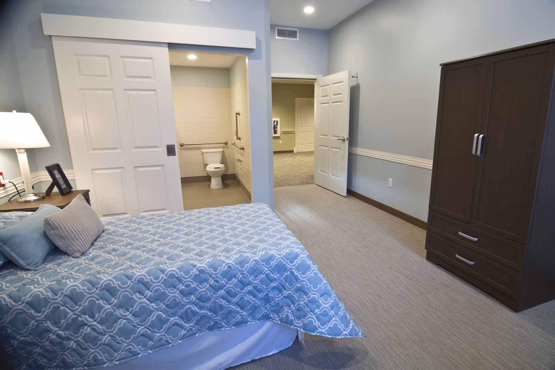 A bedroom with a bed covered in a blue patterned comforter and pillows, a bedside table with a lamp and picture frame, a dark wooden wardrobe, and an open door leading to a bathroom with a toilet and grab bars.