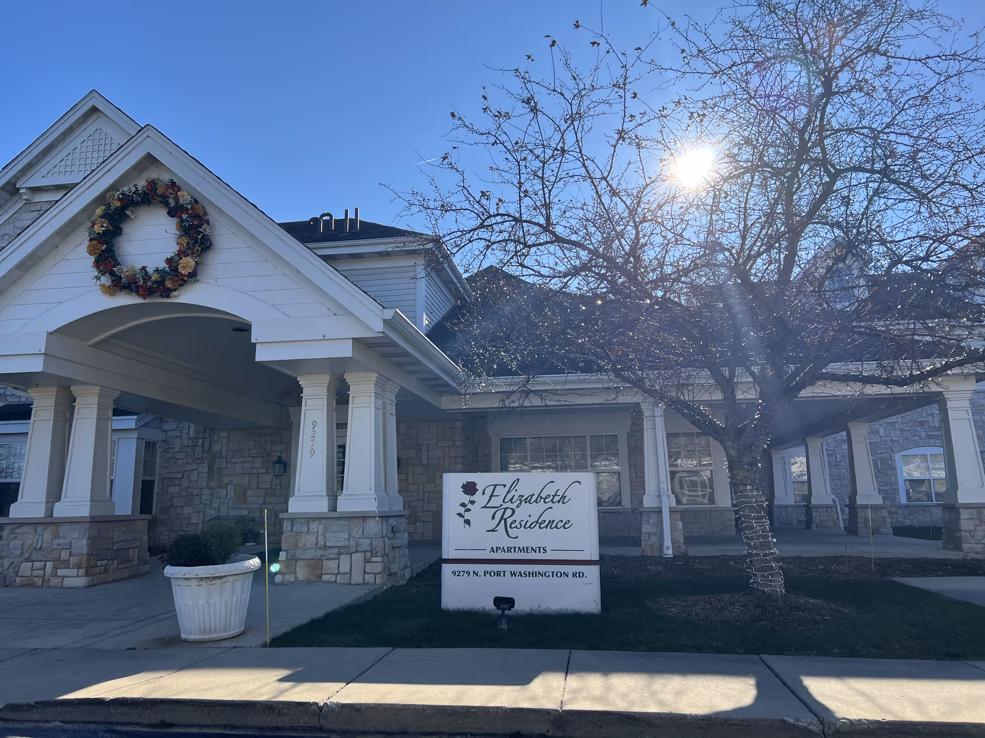 Exterior view of The Apartments At Elizabeth Residence building with stone and white siding, a covered entrance decorated with a large wreath, a tree with no leaves, and a sign displaying the facility name and address at 9279 N. Port Washington Rd.