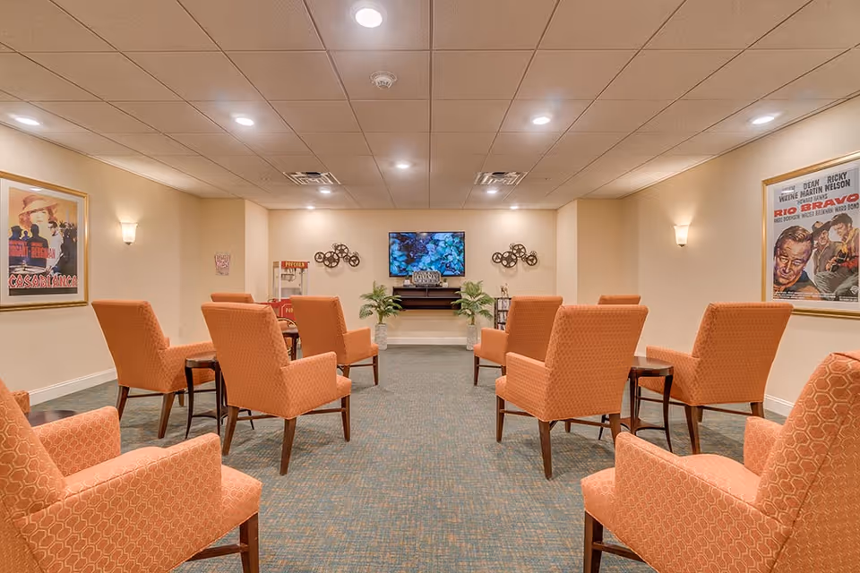 A senior living common room with rows of orange upholstered chairs facing a wall-mounted TV and framed movie posters.