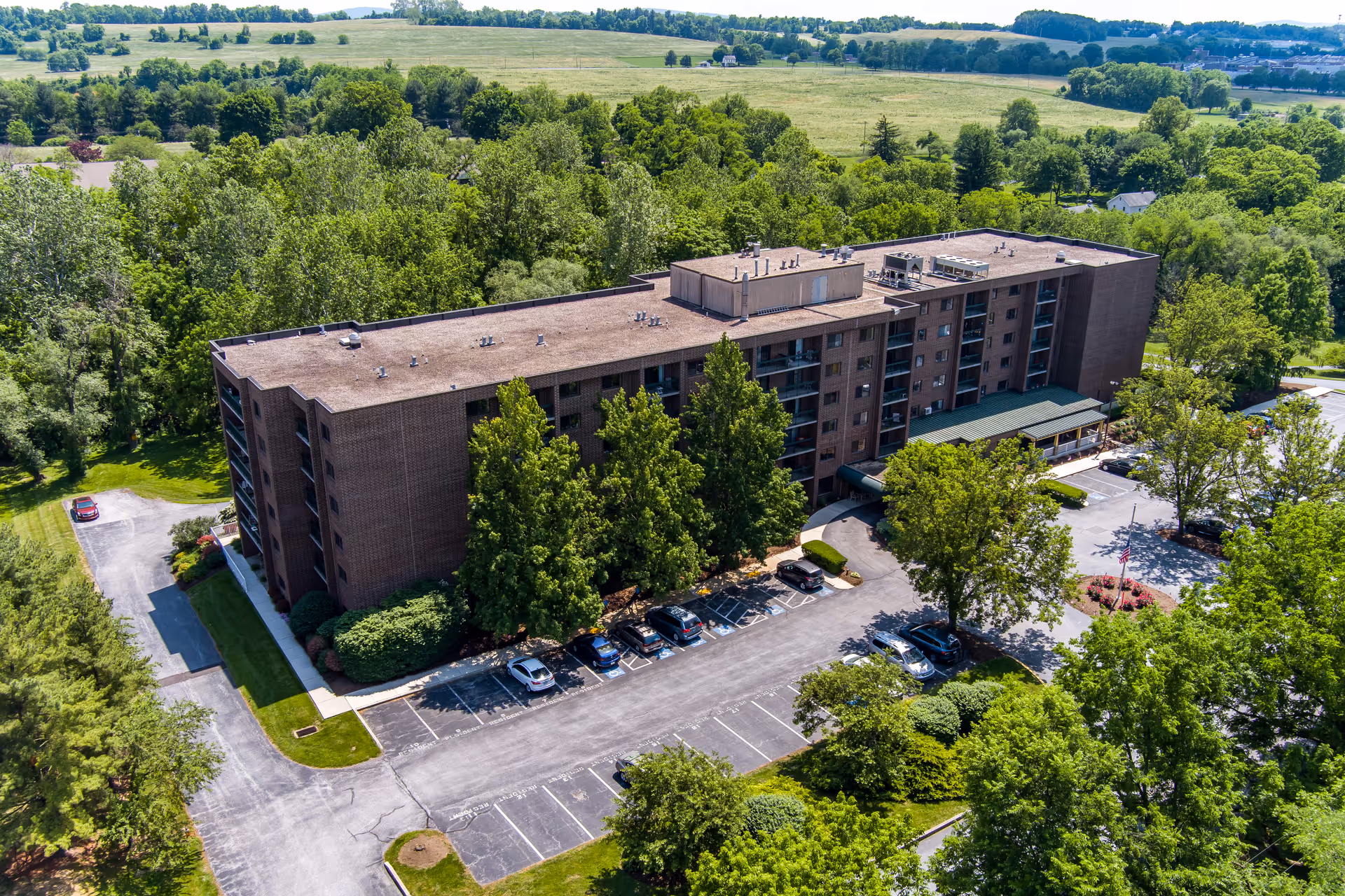Aerial view of a large multi-story brick building surrounded by trees and greenery, with a parking lot in front containing several parked cars. The building is set in a rural area with open fields and more trees in the background under a clear sky.