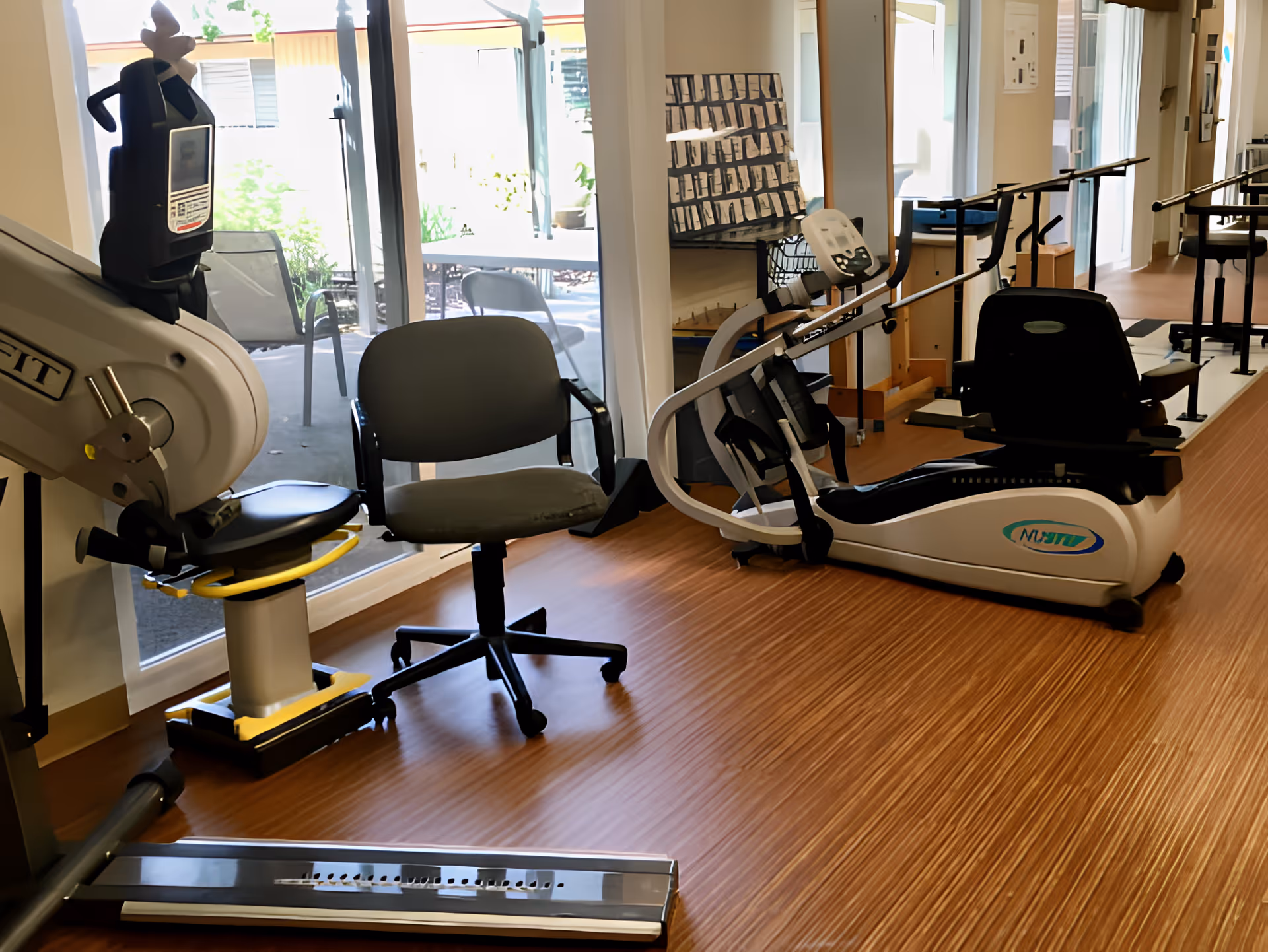 Interior view of a rehabilitation facility room with exercise equipment including a recumbent bike and parallel bars for physical therapy. There is a gray office chair and large windows showing an outdoor patio area with chairs and tables.