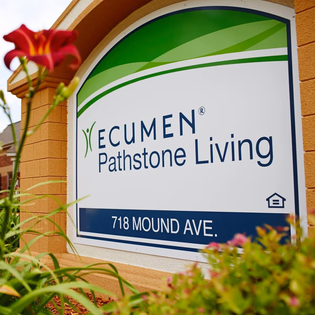 A close-up view of a sign for Ecumen Pathstone Living, located at 718 Mound Ave., surrounded by some greenery and a red flower in the foreground.