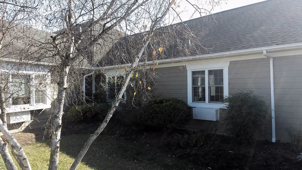 Exterior view of a single-story building with gray siding and white trim, featuring several windows and surrounded by bushes and a leafless tree in the foreground.
