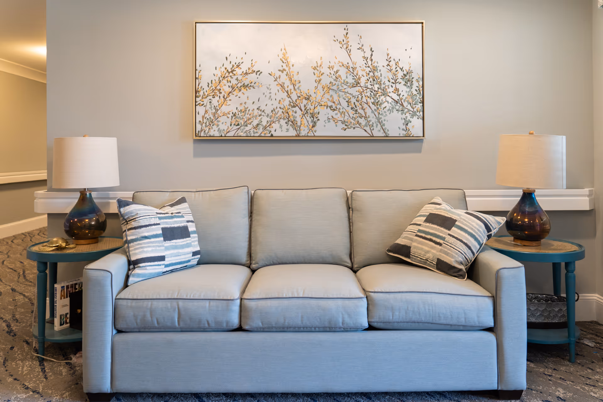 A light blue upholstered sofa with three cushions and two patterned throw pillows is centered against a light gray wall. On either side of the sofa are teal round side tables, each with a lamp featuring a dark glass base and a beige lampshade. Above the sofa hangs a large framed painting of branches with yellow and green leaves. The floor is carpeted with a patterned design.