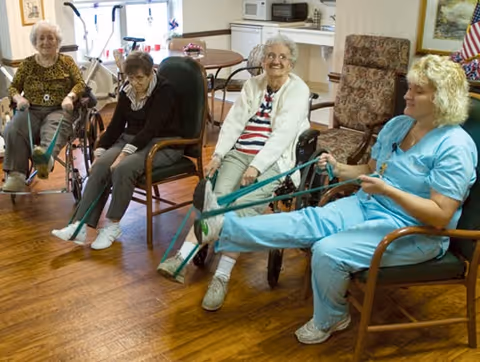 Four elderly women sitting in chairs in a common room, participating in a seated exercise using resistance bands to stretch their legs. The room has wooden flooring, a table with chairs in the background, and a kitchenette area.