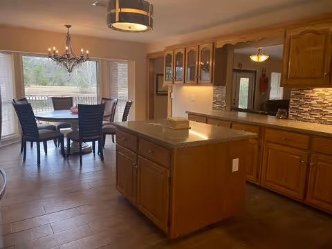 A spacious kitchen and dining area with wooden cabinets and a central island with a granite countertop. The dining area has a round table with six chairs and a chandelier above it. Large windows provide a view of the outdoor greenery.