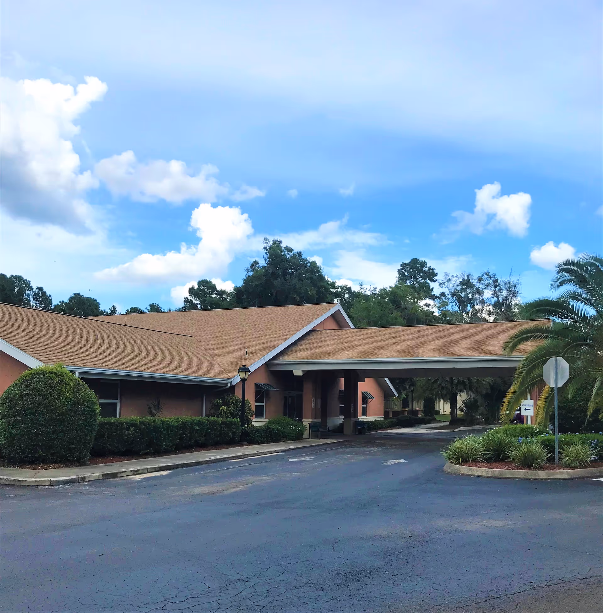 Front entrance of a single-story health and rehabilitation center with a covered drive-through canopy, landscaped shrubs and palm trees under a blue sky.
