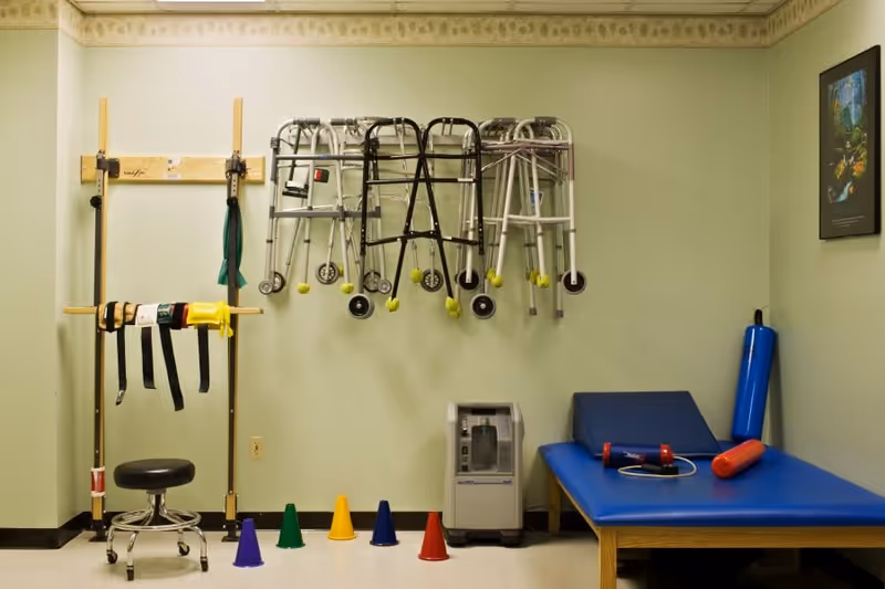 A physical therapy room with various rehabilitation equipment including walkers hanging on the wall, resistance bands on a wooden rack, a small black stool, colored cones on the floor, a blue padded therapy table with exercise equipment on it, and an oxygen concentrator machine against a light green wall.