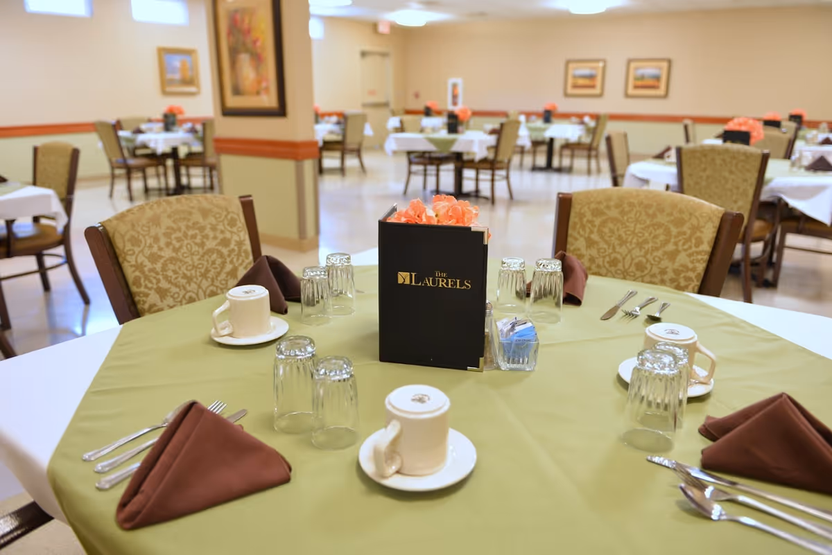 A dining room in The Laurels of Hilliard facility with round tables covered in green and white tablecloths, set with cups, glasses, silverware, and brown folded napkins. Each table has a centerpiece with a menu and orange flowers. The room has beige walls with framed artwork and multiple chairs around the tables.