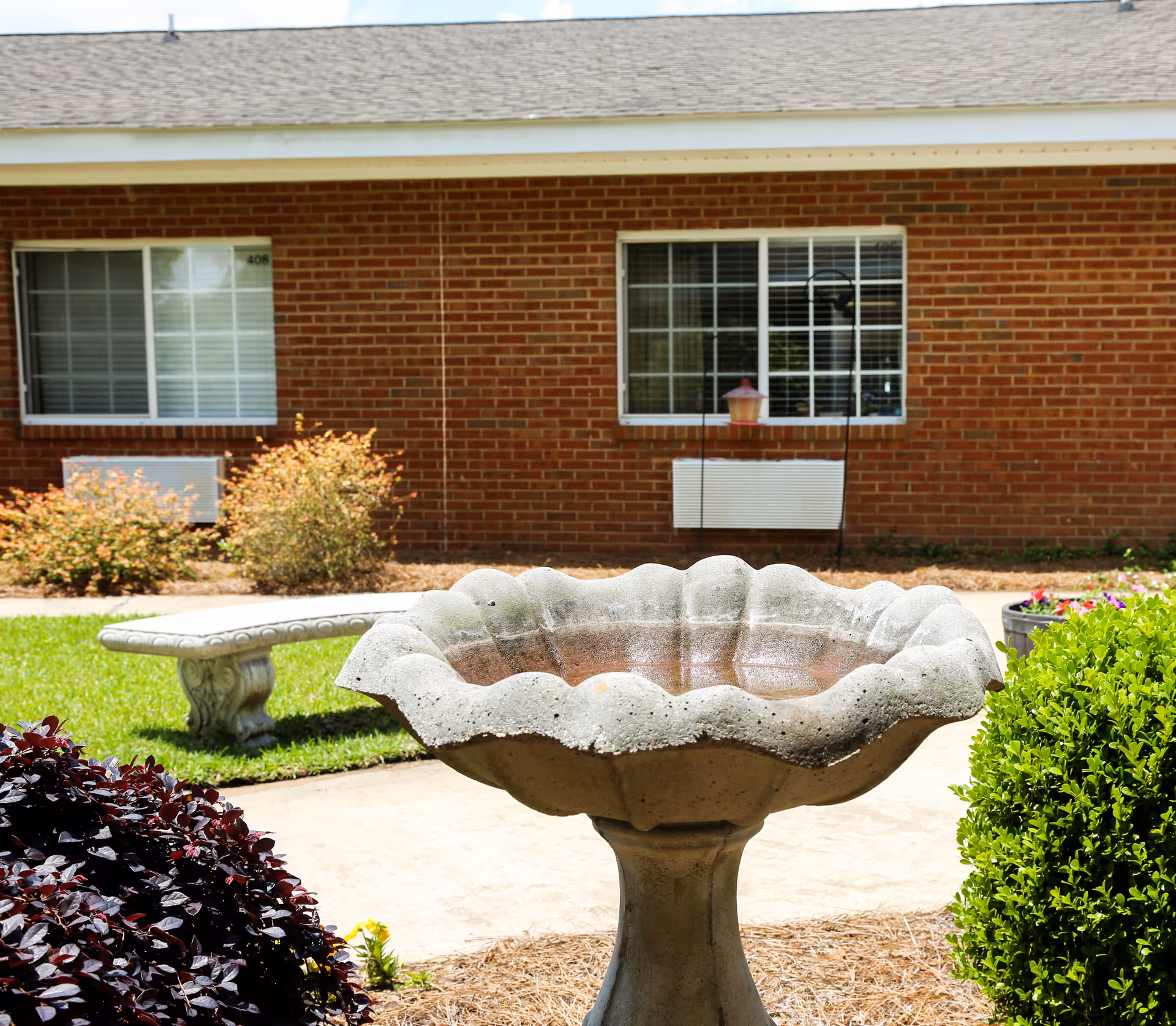 Outdoor courtyard area at Lee County Health & Rehabilitation featuring a large decorative birdbath in the foreground, a stone bench, green bushes, and a brick building with windows in the background.