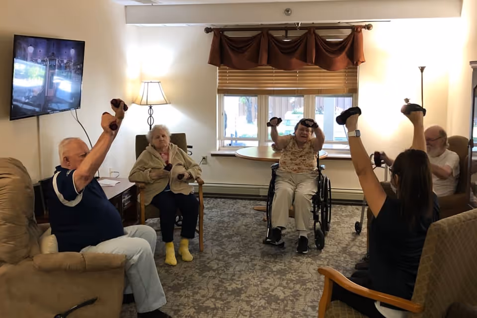 A group of elderly individuals and a caregiver participating in a seated exercise session in a well-lit living room. Some participants are holding small dumbbells and raising their arms. The room has a carpeted floor, a window with blinds and a valance, a wall-mounted TV, and several chairs.