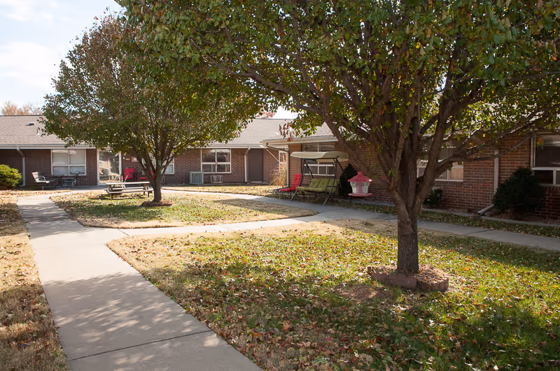 Courtyard of a single-story brick senior living facility with paved walkways, trees, and outdoor seating.
