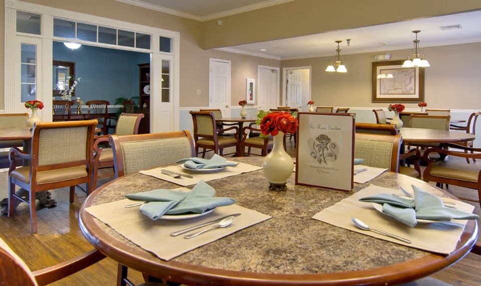 A dining room in a senior living facility with multiple round tables set with placemats, folded napkins, and silverware. Each table has a small vase with red flowers and a menu card. The room has wooden flooring, beige walls, and overhead lighting fixtures. There is an adjacent room visible through a large doorway with additional seating and a blue accent wall.