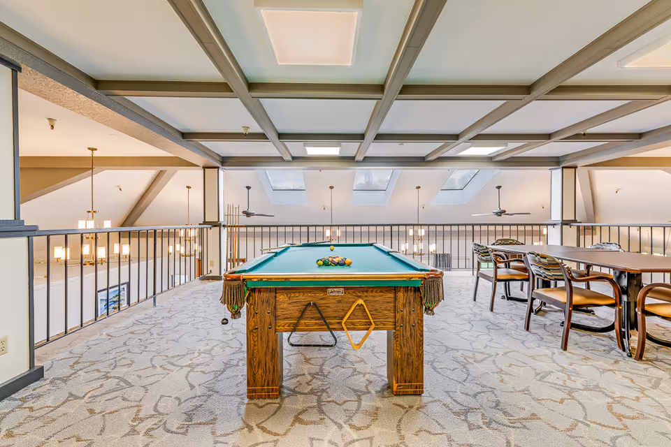 Interior view of a senior living facility's recreational area featuring a pool table with balls arranged for a game. The room has a patterned carpet, a coffered ceiling with skylights, ceiling fans, and hanging light fixtures. To the right, there is a long table with several chairs around it. The area is bordered by a railing overlooking a lower level.