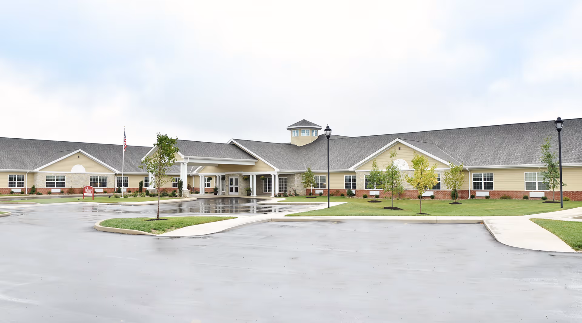 Front exterior of a low, single-story senior living building with a covered entrance, flagpole, landscaping, and a wet parking lot.