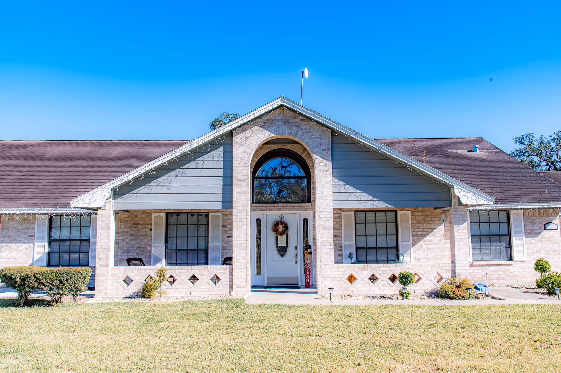 Front exterior view of a single-story brick building with a peaked roof and a central entrance featuring an arched window above the door. The building has large windows with white shutters and a well-maintained lawn with small bushes in front. The sky is clear and blue.
