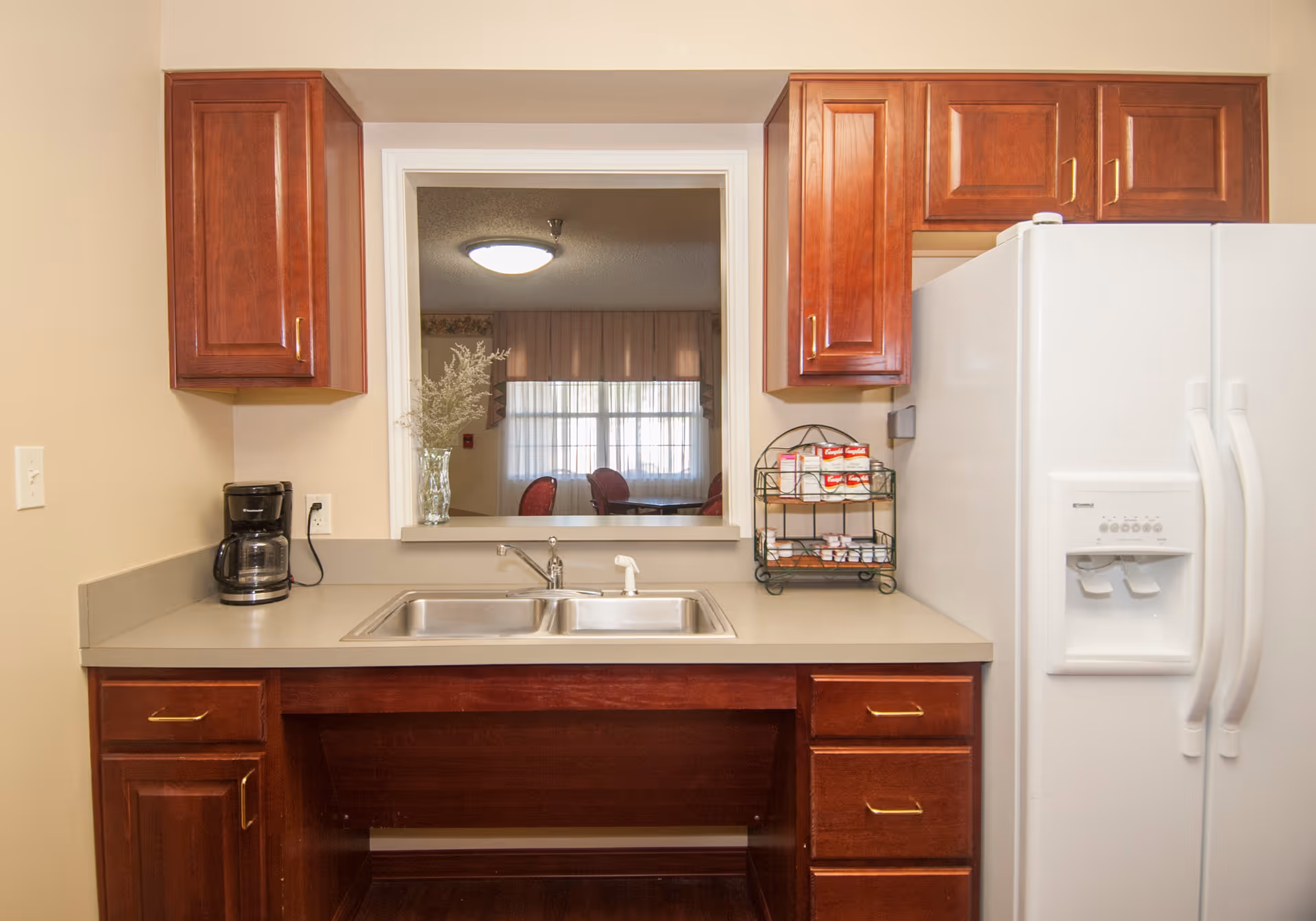 A kitchen area with wooden cabinets, a double stainless steel sink, a coffee maker on the left countertop, and a white refrigerator with a water and ice dispenser on the right. There is a small rack with coffee supplies on the counter next to the refrigerator. A window above the sink looks into another room with a round table and red chairs.