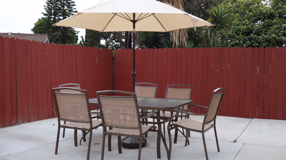 Outdoor patio area with a glass-top table surrounded by six brown metal chairs with mesh seats and backs. A large beige umbrella is positioned in the center of the table. The patio is enclosed by a red wooden fence, and there are green trees and bushes visible behind the fence.