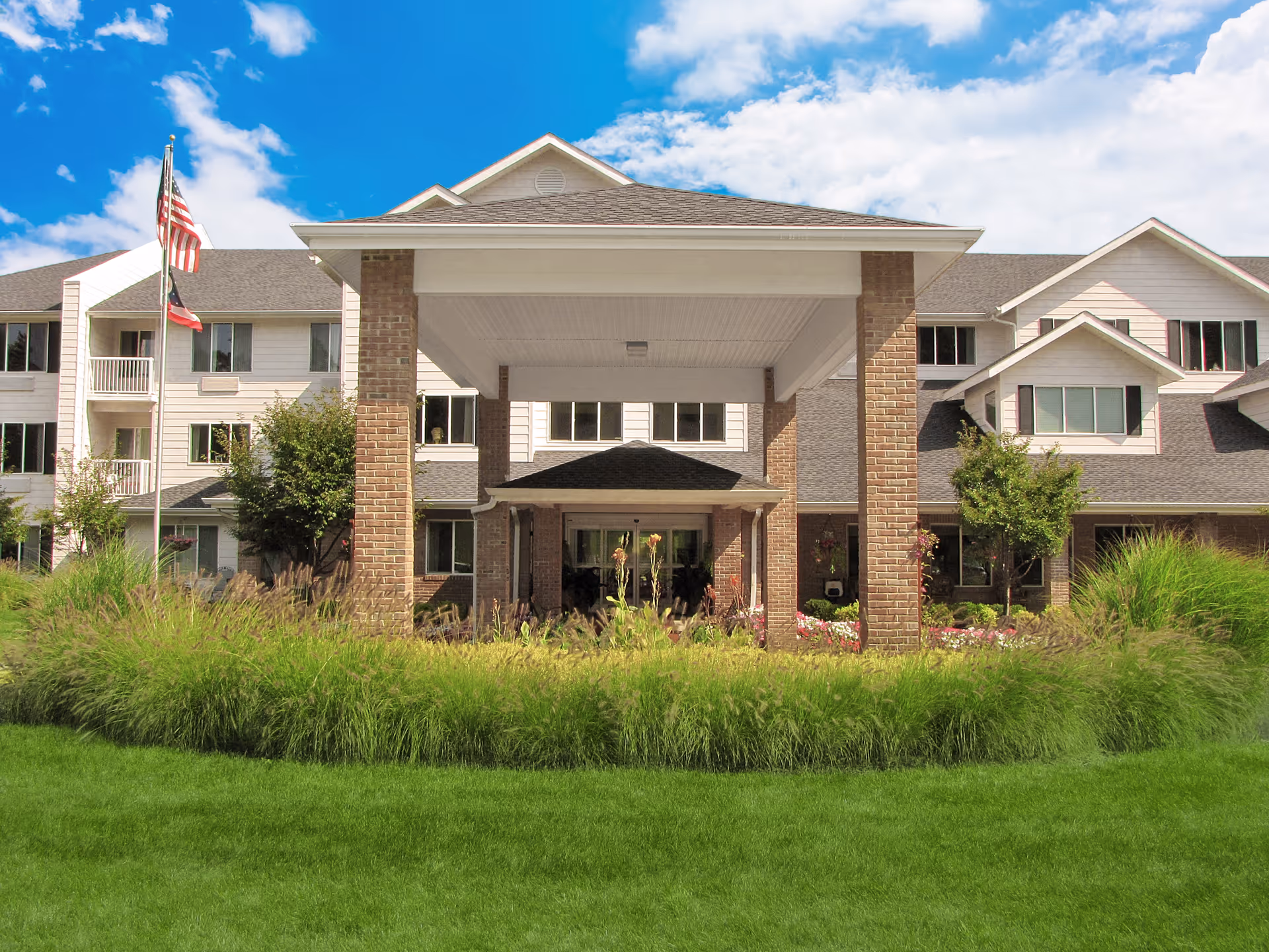 Front exterior view of a multi-story senior living facility with a covered entrance supported by brick columns, surrounded by well-maintained green grass and landscaping, under a partly cloudy blue sky.