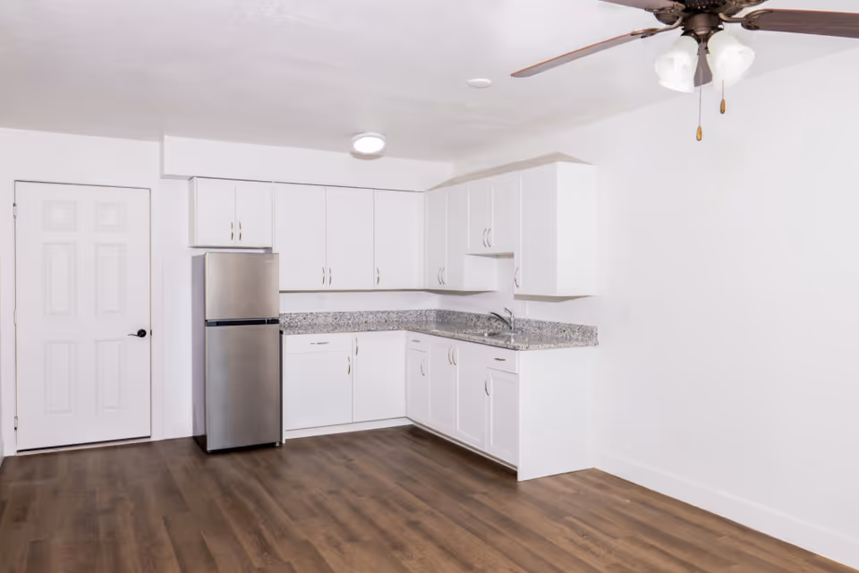 A clean, modern kitchen area with white cabinets, granite countertops, a stainless steel refrigerator, and a ceiling fan with lights. The floor is wood, and there is a white door to the left.