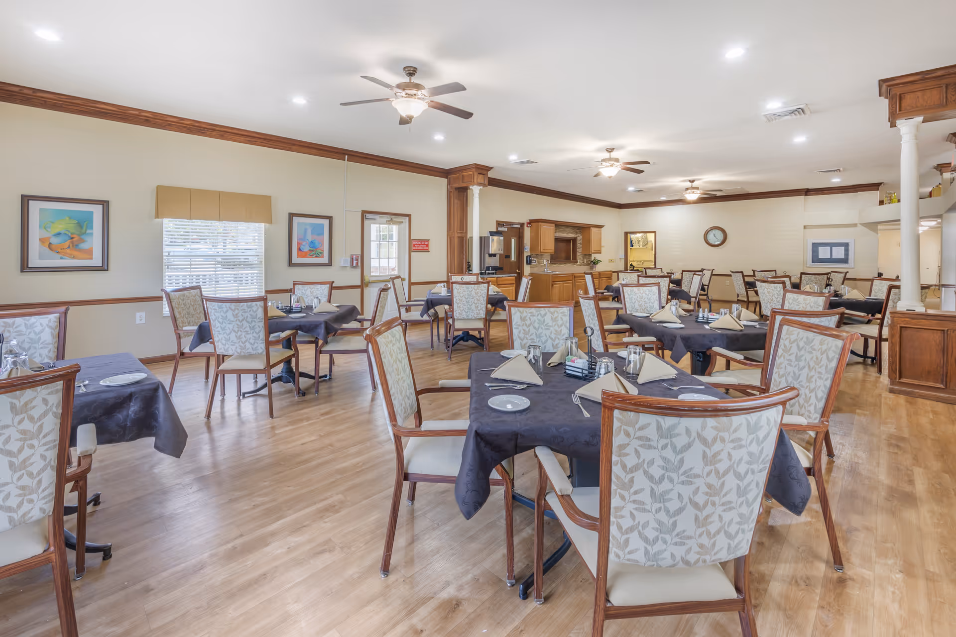 A spacious dining room with multiple tables covered in dark tablecloths and set with napkins, plates, glasses, and silverware. The chairs have wooden frames and patterned upholstery. The room has light-colored walls with wooden trim, ceiling fans with lights, and framed artwork on the walls. There is a serving area with wooden cabinets and a clock on the far wall.