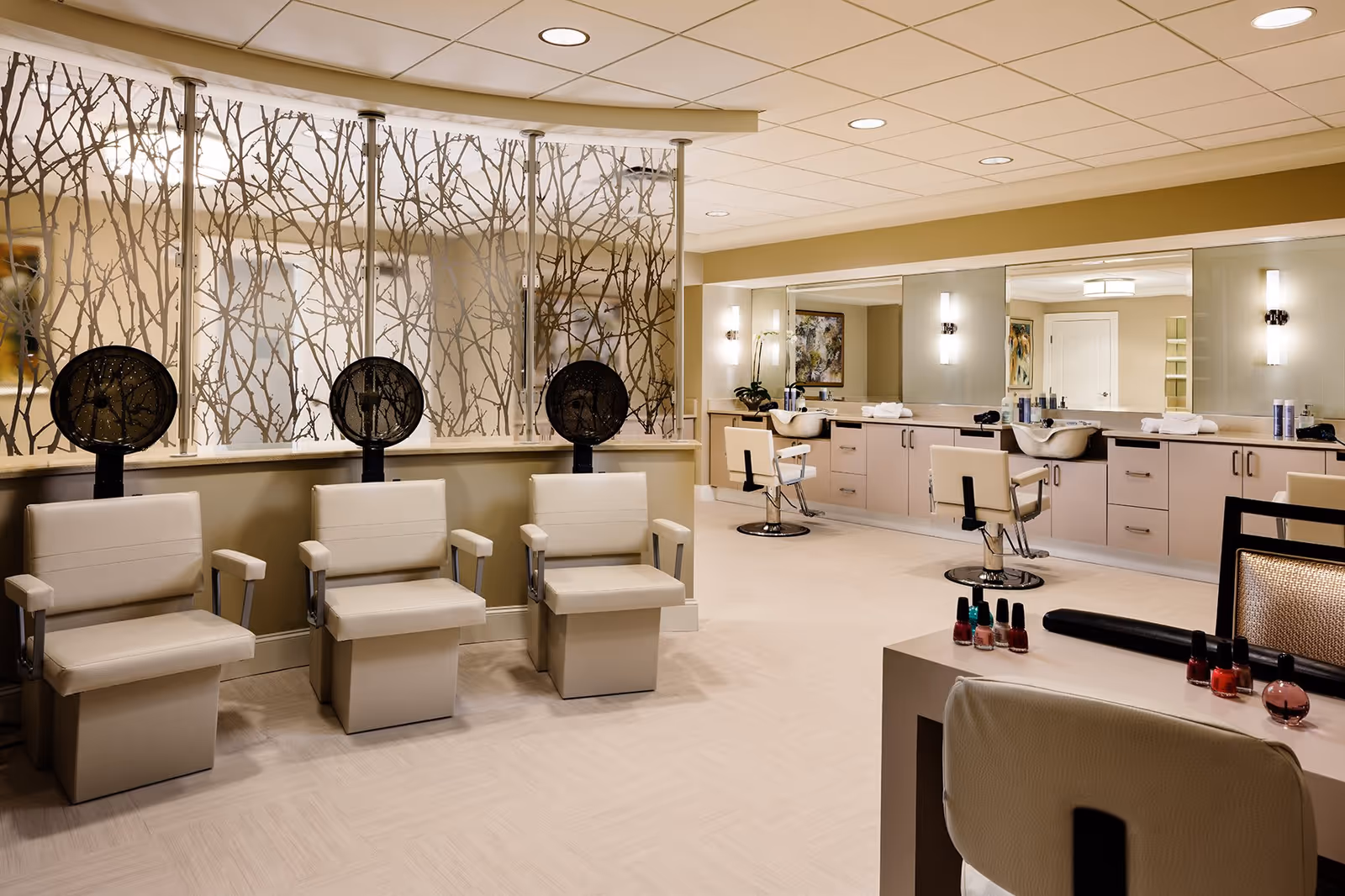 A modern salon area in a senior living facility featuring three beige salon chairs with hair dryers, a counter with two styling chairs and sinks, large mirrors, and a table with various nail polish bottles. The room has warm lighting and decorative glass panels with a branch pattern.