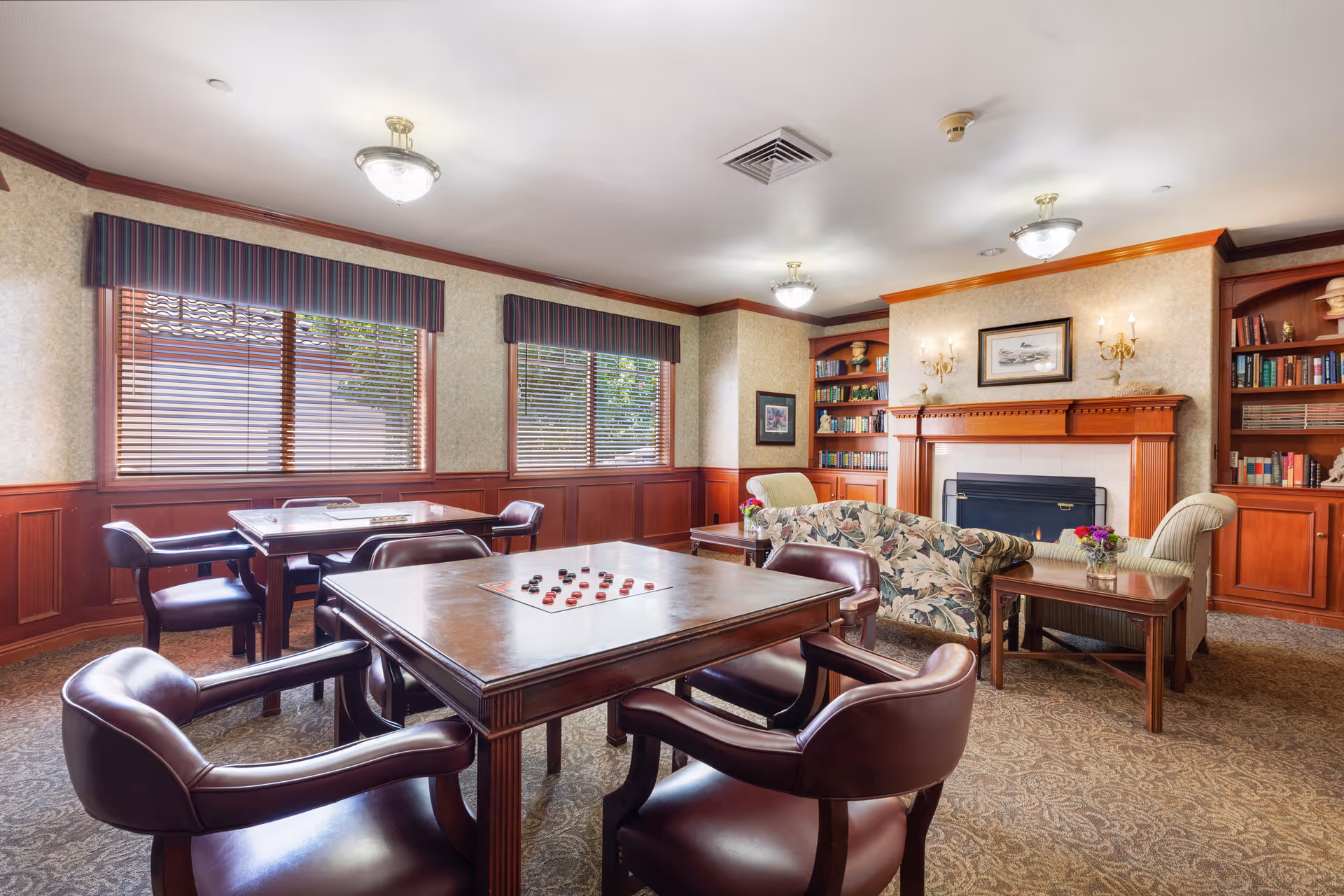A cozy common room in a senior living facility featuring two wooden tables with leather chairs arranged around them. One table has a checkers game set up. The room has large windows with striped valances, wood paneling on the lower walls, and built-in bookshelves filled with books. There is a floral-patterned sofa and two armchairs positioned around a wooden coffee table in front of a fireplace with a wooden mantel, decorated with a framed picture and wall sconces.