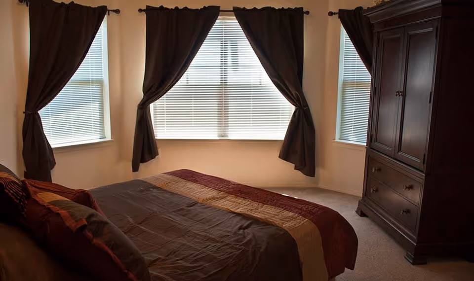 Bedroom with a bed dressed in a multi-colored quilt, three windows with blinds and dark curtains, and a large wooden armoire.