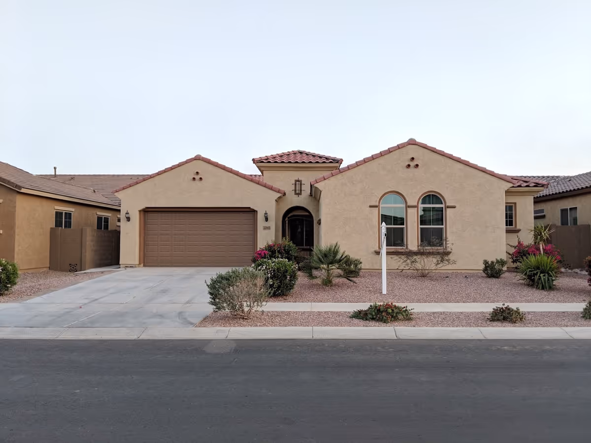 Front exterior view of a single-story house with a two-car garage, beige stucco walls, red tile roof, and desert landscaping with bushes and small plants.