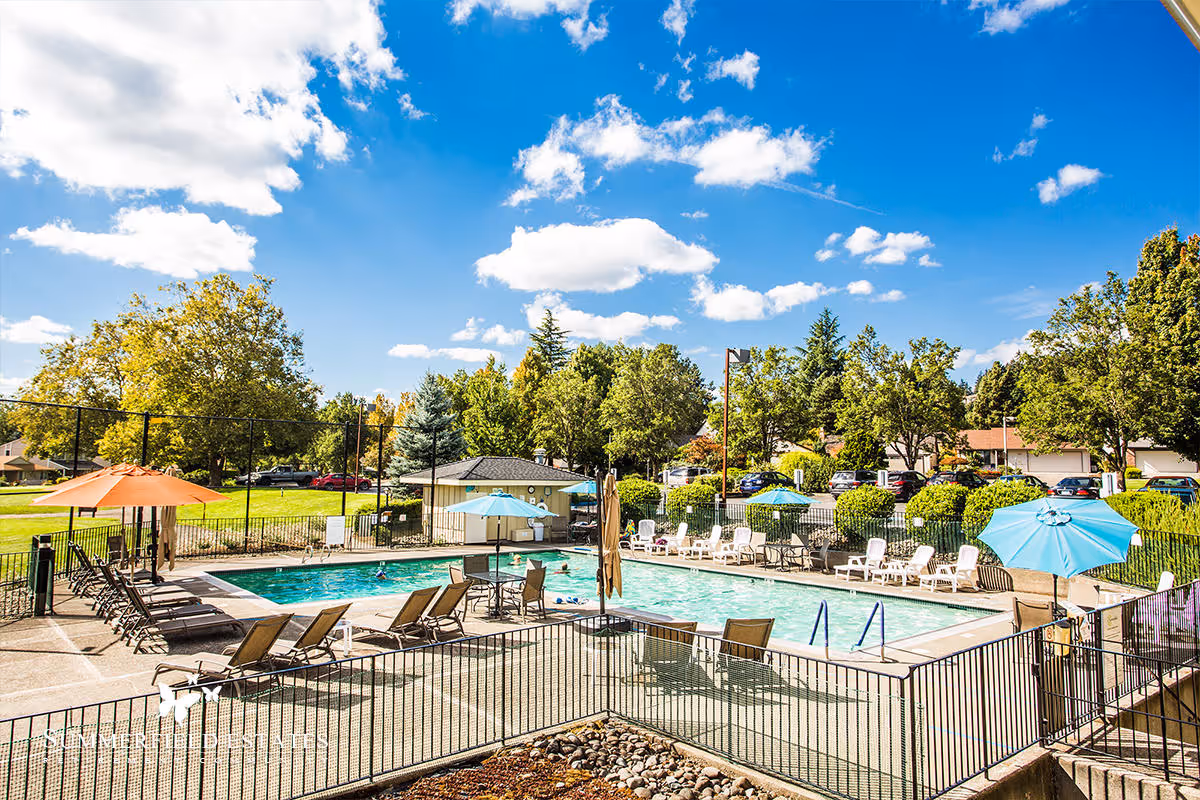 Outdoor swimming pool area at Summerfield Estates Retirement Community with lounge chairs, tables with umbrellas, and a fenced perimeter. The pool is surrounded by trees, greenery, and a parking lot with cars under a partly cloudy blue sky.