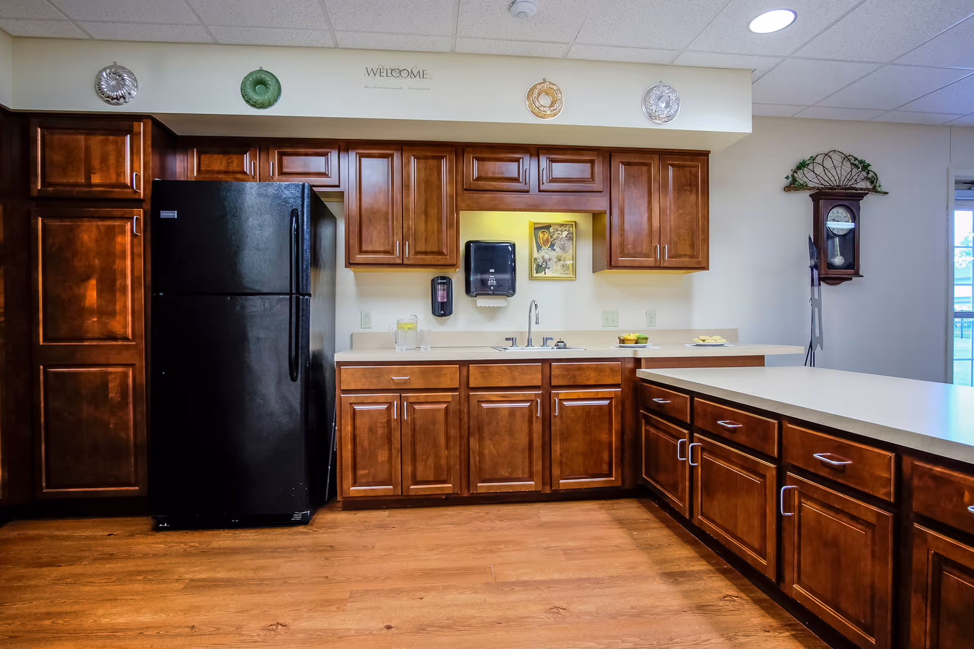 A kitchen area with wooden cabinets and drawers, a black refrigerator, a sink with a faucet, and a countertop with a glass pitcher of water and a plate of cookies. Decorative plates and a 'WELCOME' sign are displayed above the cabinets. A wall clock is mounted on the right wall near a window.
