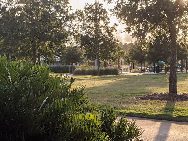 A sunlit outdoor park area with green grass, trees, and bushes. There are a few people visible in the distance near a playground structure. The sky is partly cloudy with sunlight filtering through the trees.