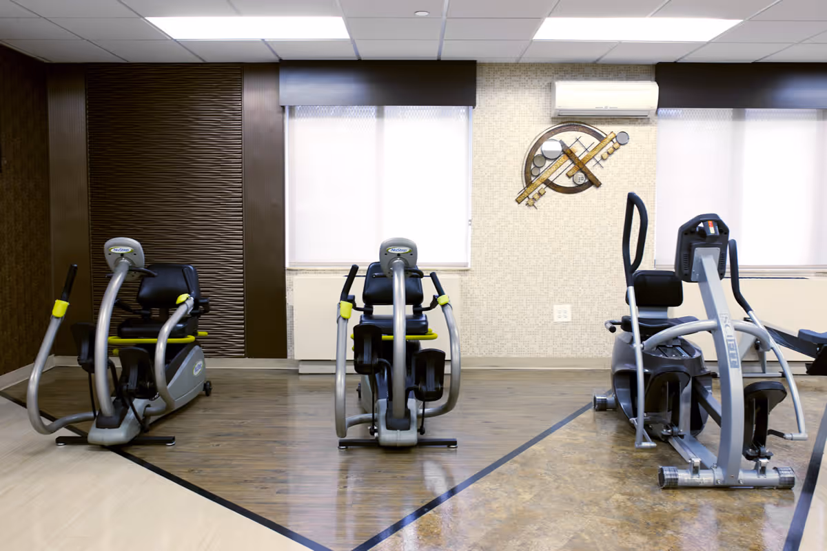 Indoor exercise room with three pieces of cardio equipment, including two recumbent bikes and one elliptical machine, set against a wall with two windows covered by white blinds and a decorative wall art piece. The room has a mix of wood and tile flooring and a white ceiling with fluorescent lighting.