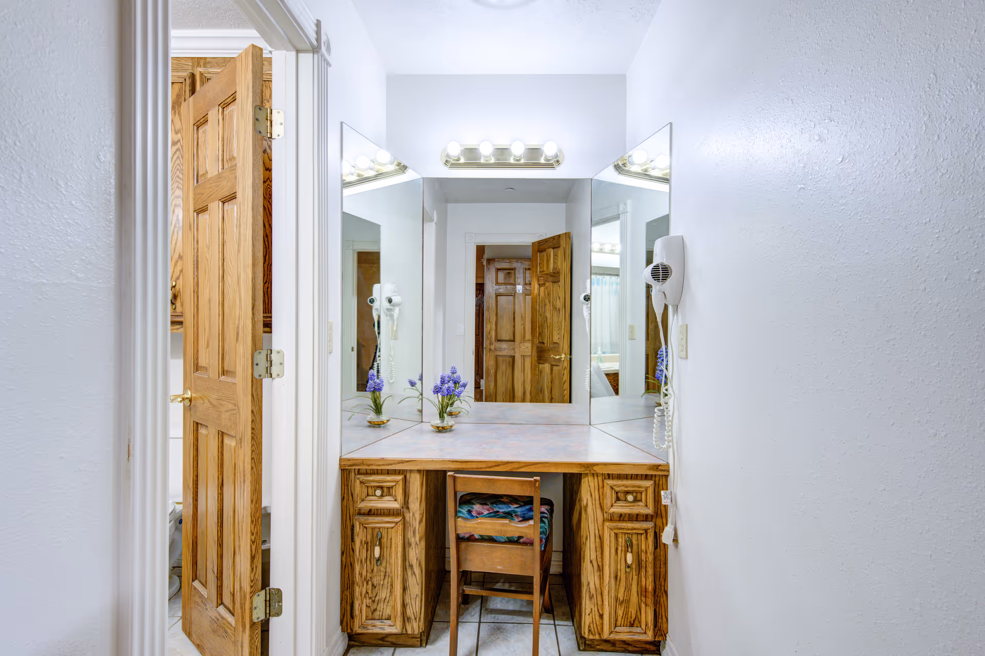 A vanity area with a large mirror and wooden cabinetry, featuring a small wooden chair with a colorful cushion. To the left, there is an open wooden door leading to a bathroom with a visible toilet. The walls are white, and a wall-mounted hairdryer is attached to the right side of the vanity.