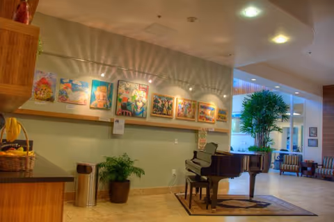 Interior view of a senior living facility featuring a black grand piano on a rug, a row of colorful paintings displayed on the wall above a ledge, a potted plant next to a trash can, and seating areas with chairs and large windows in the background.
