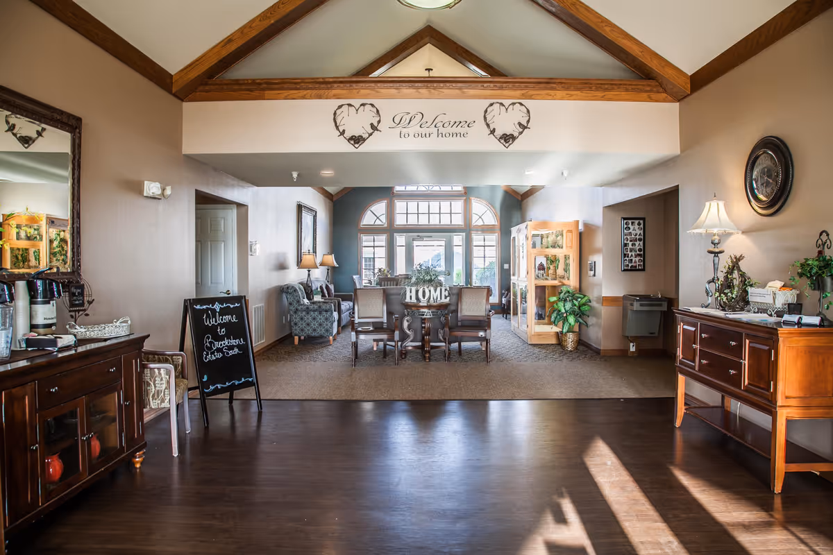 A warmly lit interior common area with wooden beams on the ceiling and a welcoming sign that reads 'Welcome to our home'. The room features a seating area with chairs and a table, a chalkboard sign that says 'Welcome to Brookstone Estates South', decorative plants, a lamp on a wooden sideboard, and large windows letting in natural light.