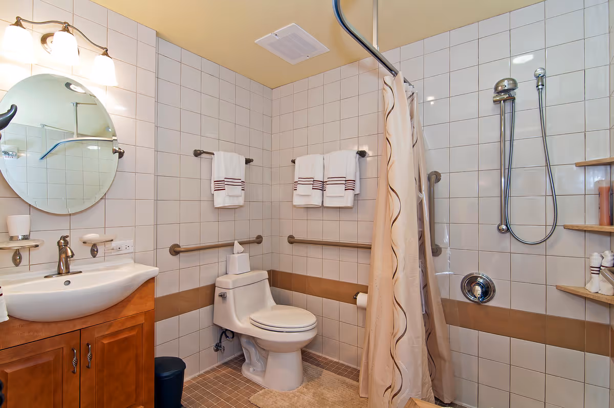 Accessible tiled bathroom with a toilet, sink vanity and round mirror, grab bars, towels, and a shower with curtain.