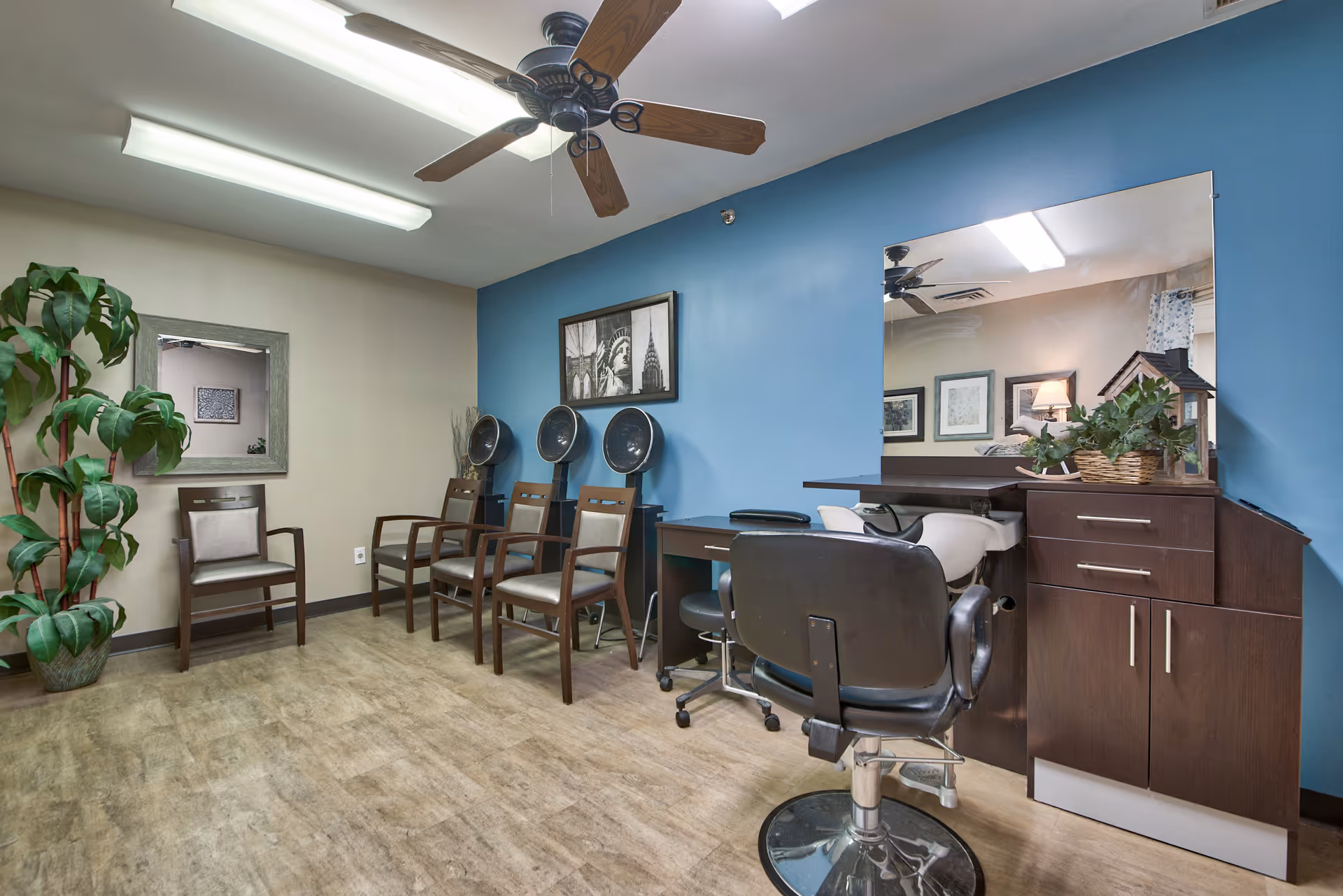 Interior view of a salon area in a senior living facility with three hair drying chairs against a blue wall, a large mirror above a dark wood counter with a salon chair in front, additional chairs along the opposite wall, a ceiling fan, and a potted plant in the corner.