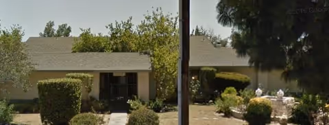 Single-story beige building with a pitched roof, central walkway and landscaped shrubs and trees in the front yard.