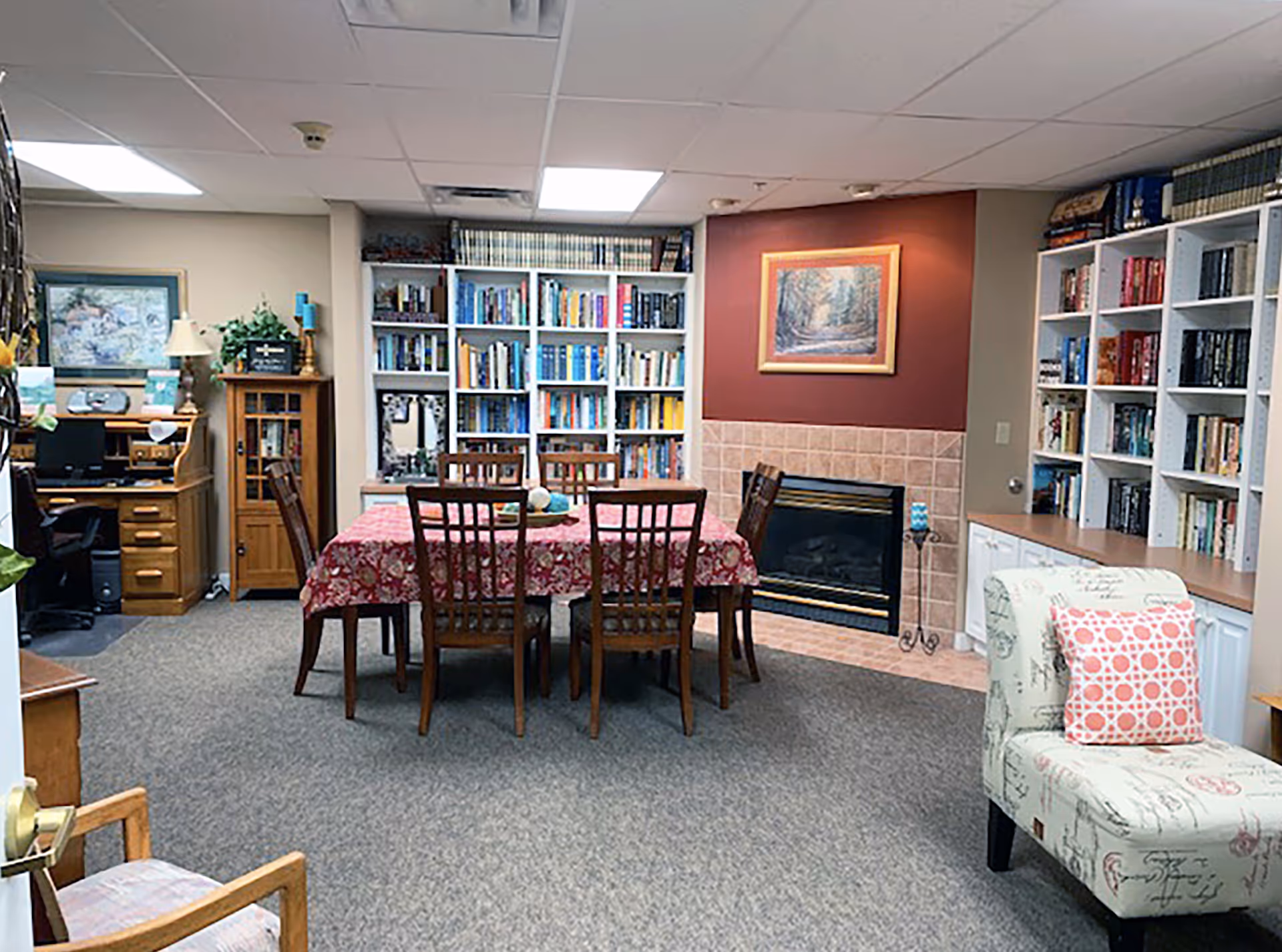 A cozy room in Peachtree Village Retirement Community featuring a wooden dining table with six chairs covered by a red floral tablecloth. Behind the table is a fireplace with a framed painting above it, flanked by white bookshelves filled with books. To the left, there is a wooden desk with a computer and a small cabinet with a lamp and decorative items. On the right side, there is a cushioned chair with a pink patterned pillow. The room has carpeted flooring and a drop ceiling with fluorescent lights.