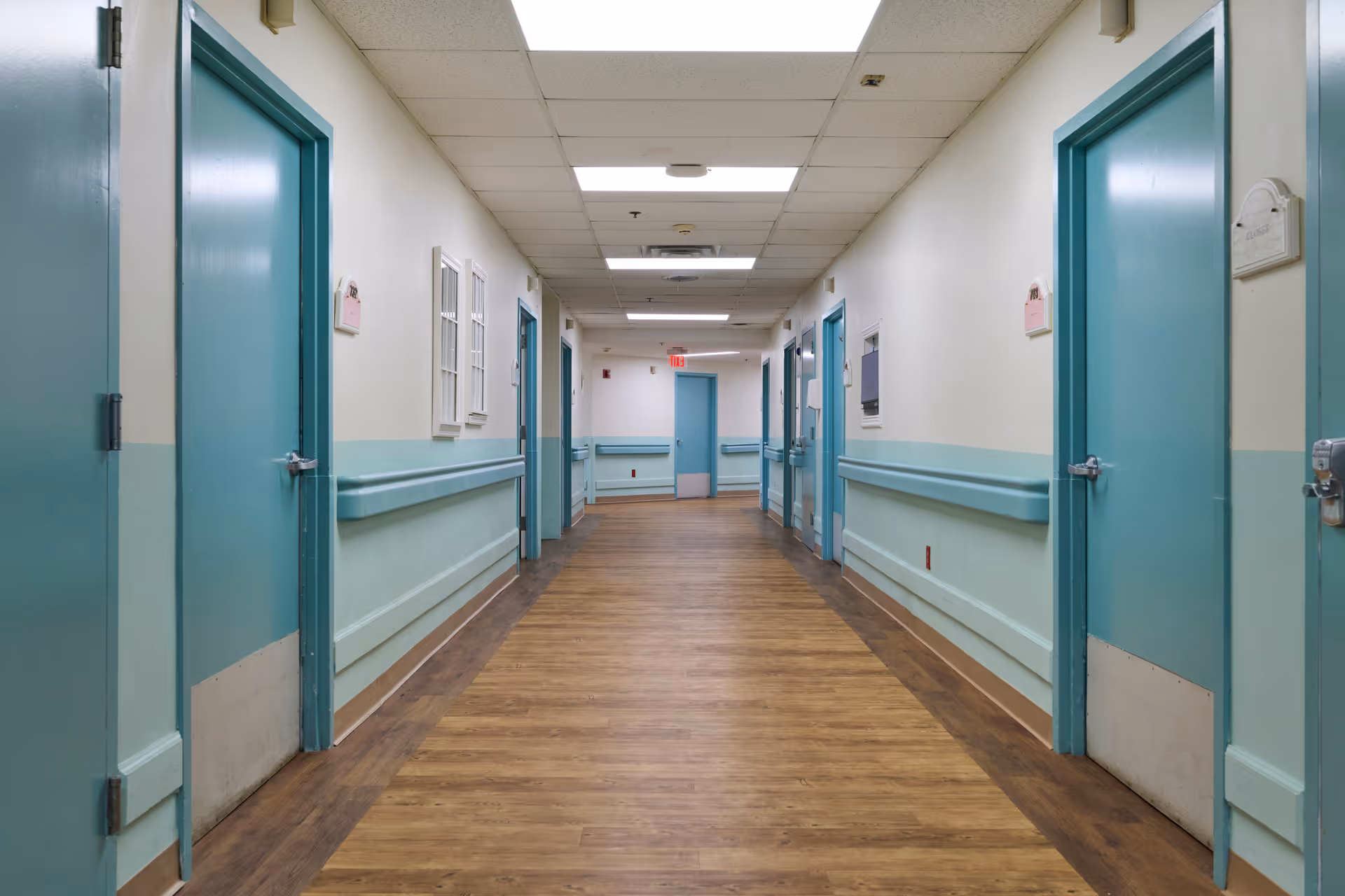 Long empty corridor with teal doors, handrails, and wood-look flooring in a healthcare facility.