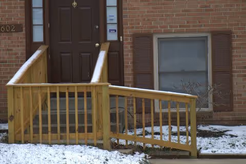 Entrance to a brick building with a brown door and a wooden ramp leading up to it. There is a window with brown shutters to the right of the door, and snow covers the ground around the ramp and building.