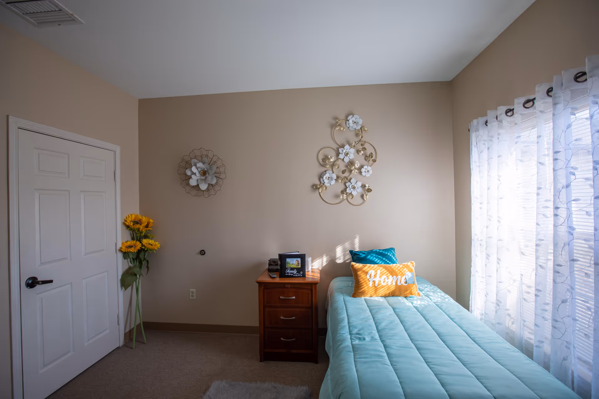 A small bedroom with a single bed covered in a light blue comforter and two pillows, one teal and one yellow with the word 'Home' on it. Next to the bed is a wooden nightstand with a small picture frame and a decorative item. The walls are beige with two metal floral wall decorations. A white door is on the left side, and a tall vase with sunflowers stands beside it. Sheer white curtains cover a window on the right, allowing sunlight to softly illuminate the room.