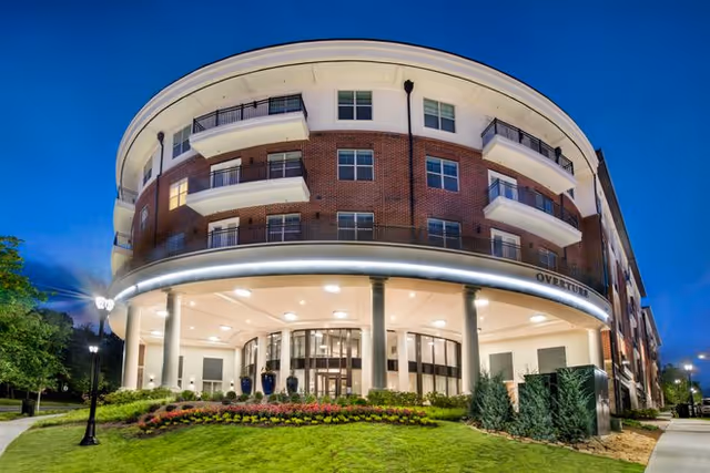 Exterior view of a multi-story senior living facility named Overture Buckhead South at dusk, featuring a curved facade with balconies, large windows, well-lit entrance with columns, landscaped garden with flowers and shrubs, and a clear evening sky.