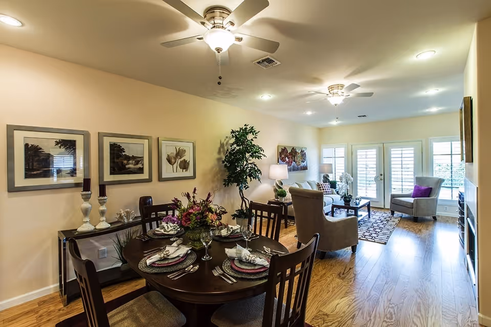 A well-lit living and dining area in a senior living facility. The dining area features a round wooden table set with plates, napkins, and a floral centerpiece. The living area has comfortable armchairs, a sofa, a coffee table, and large windows with white shutters letting in natural light. The walls are decorated with framed artwork and there are ceiling fans with lights.