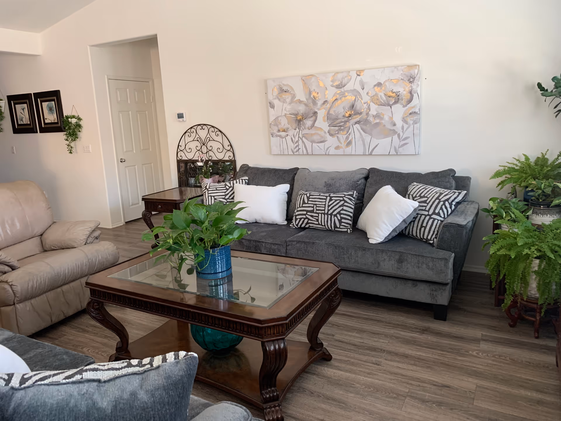 A cozy living room with a gray sofa adorned with patterned and white pillows, a glass-top wooden coffee table with a green potted plant, a beige recliner chair, wall art featuring flowers, and several green plants placed around the room. The floor is wooden, and there is a white door and framed pictures on the wall.