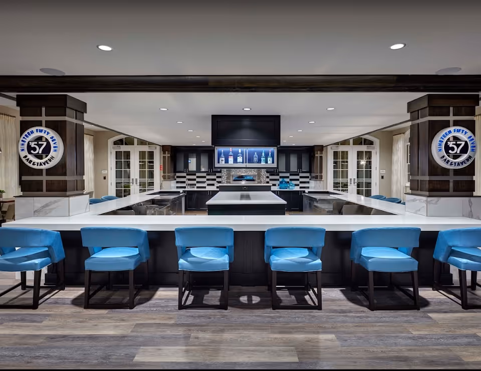 Modern bar area with a white countertop and six blue cushioned bar stools. Behind the bar is a kitchen setup with black and white cabinetry, a central island, and a display of liquor bottles on a backlit shelf. The space features wood accents and two pillars with circular signs that read 'Nineteen Fifty Seven Bar & Tavern'.