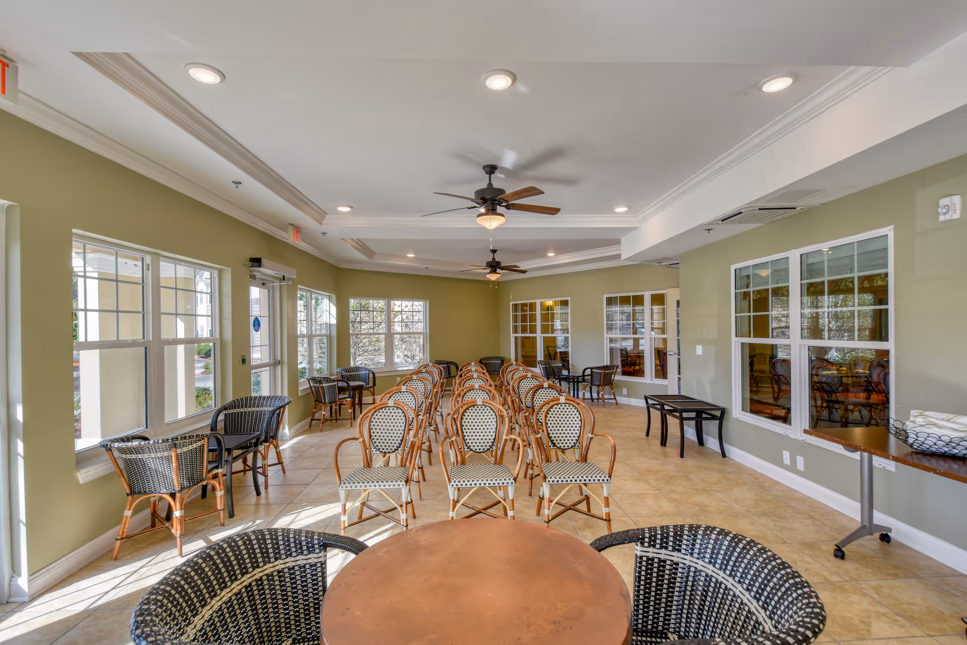 A spacious room with rows of chairs arranged facing forward, several small tables with wicker chairs near the windows, ceiling fans, and large windows letting in natural light.
